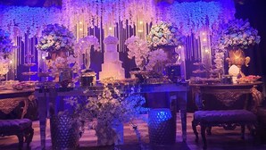 Ein Innenraum mit hoher Decke, verziert mit hängenden floralen Arrangements und einer mehrstufigen Torte in der Mitte. Die Tische sind mit floralen Mittelstücken und Leuchten gedeckt.