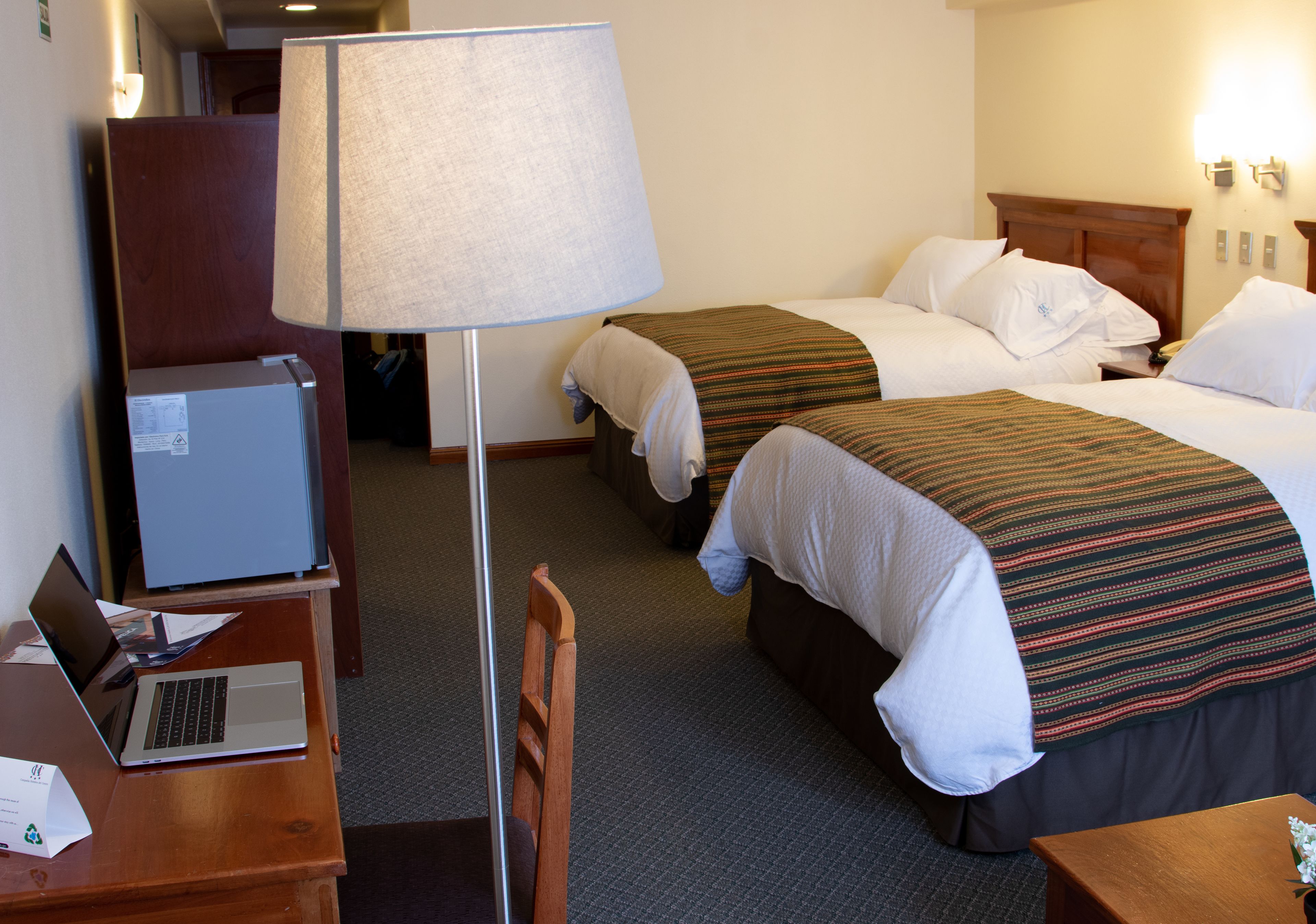 A hotel room with two beds, a desk with a laptop, a chair, and a lamp.