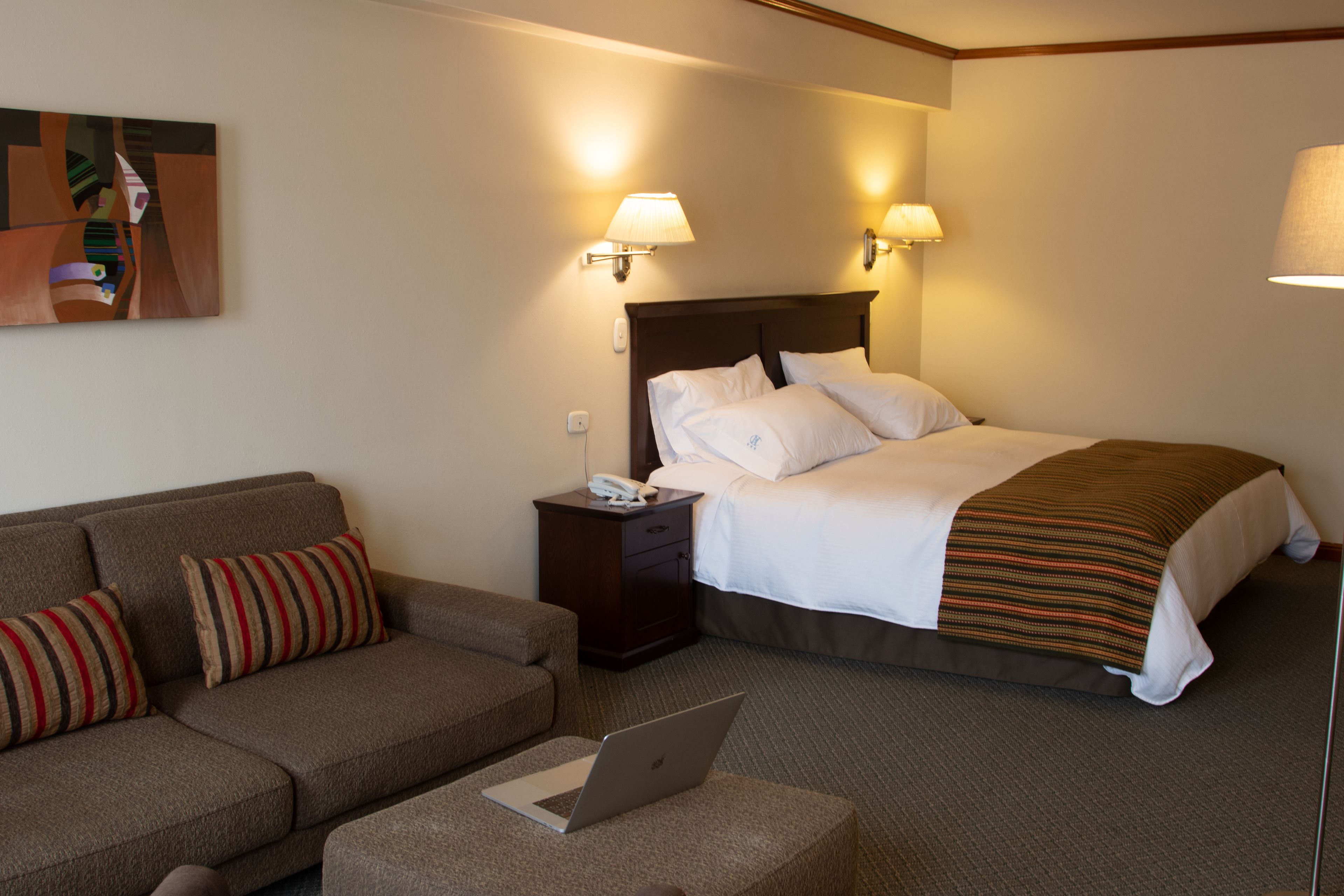 A hotel room with a bed, a sofa, a nightstand with a phone, and a laptop on a small table.