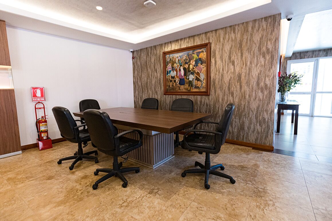 A conference room with a large table, four chairs, a fire extinguisher, and a painting on the wall.