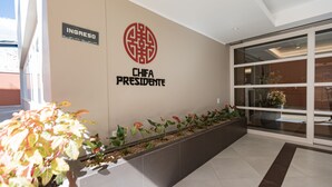 Zona de recepción con un cartel que indica “INGRESO” y “CHIFA PRESIDENTE” en la pared, una maceta con plantas y una puerta de vidrio que da al interior.