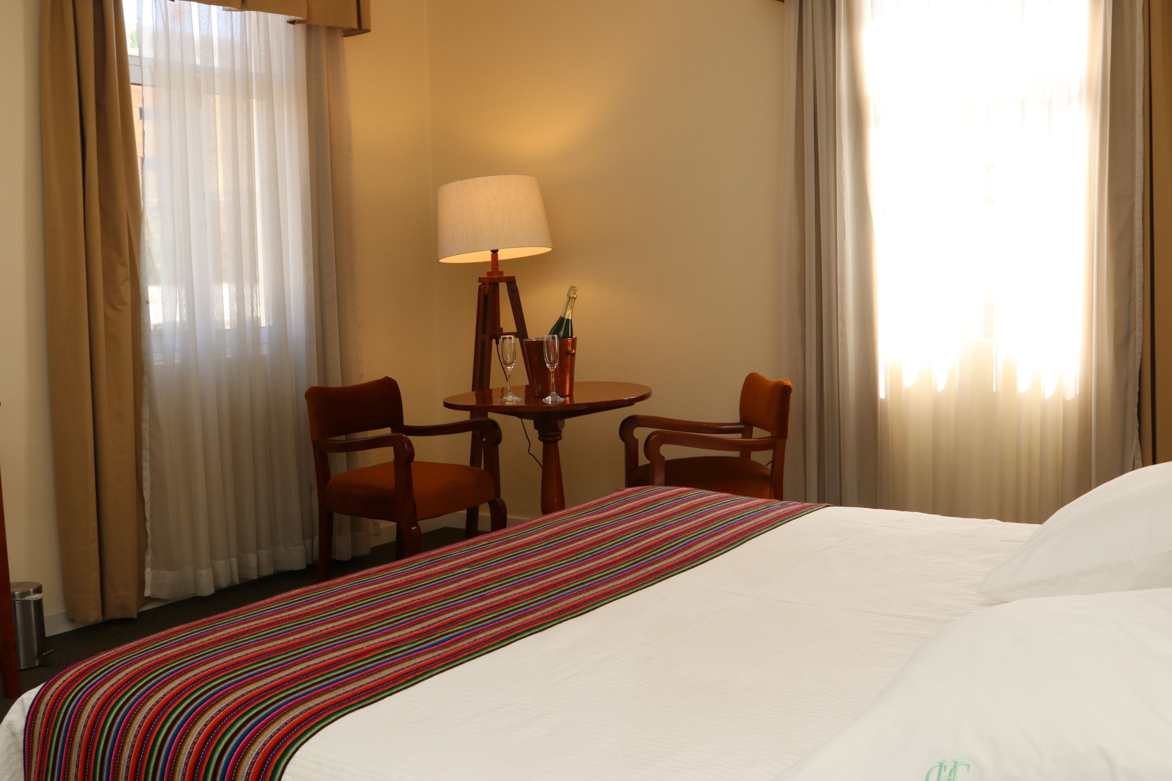 Une chambre d’hôtel avec un lit, une petite table avec une lampe, deux chaises et une fenêtre avec des rideaux.