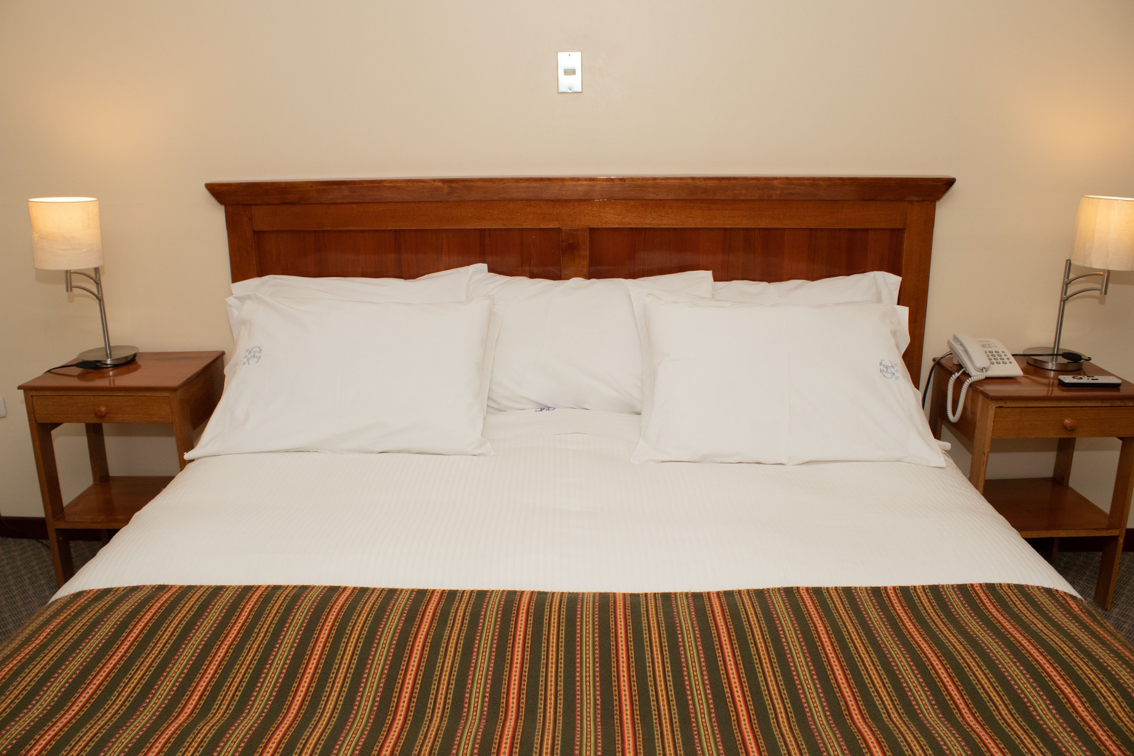 Un lit bien fait, avec du linge de lit blanc, une tête de lit en bois et une couvre-lit à rayures. Il y a deux tables de chevet, chacune avec une lampe, et un téléphone est posé sur l’une d’elles.