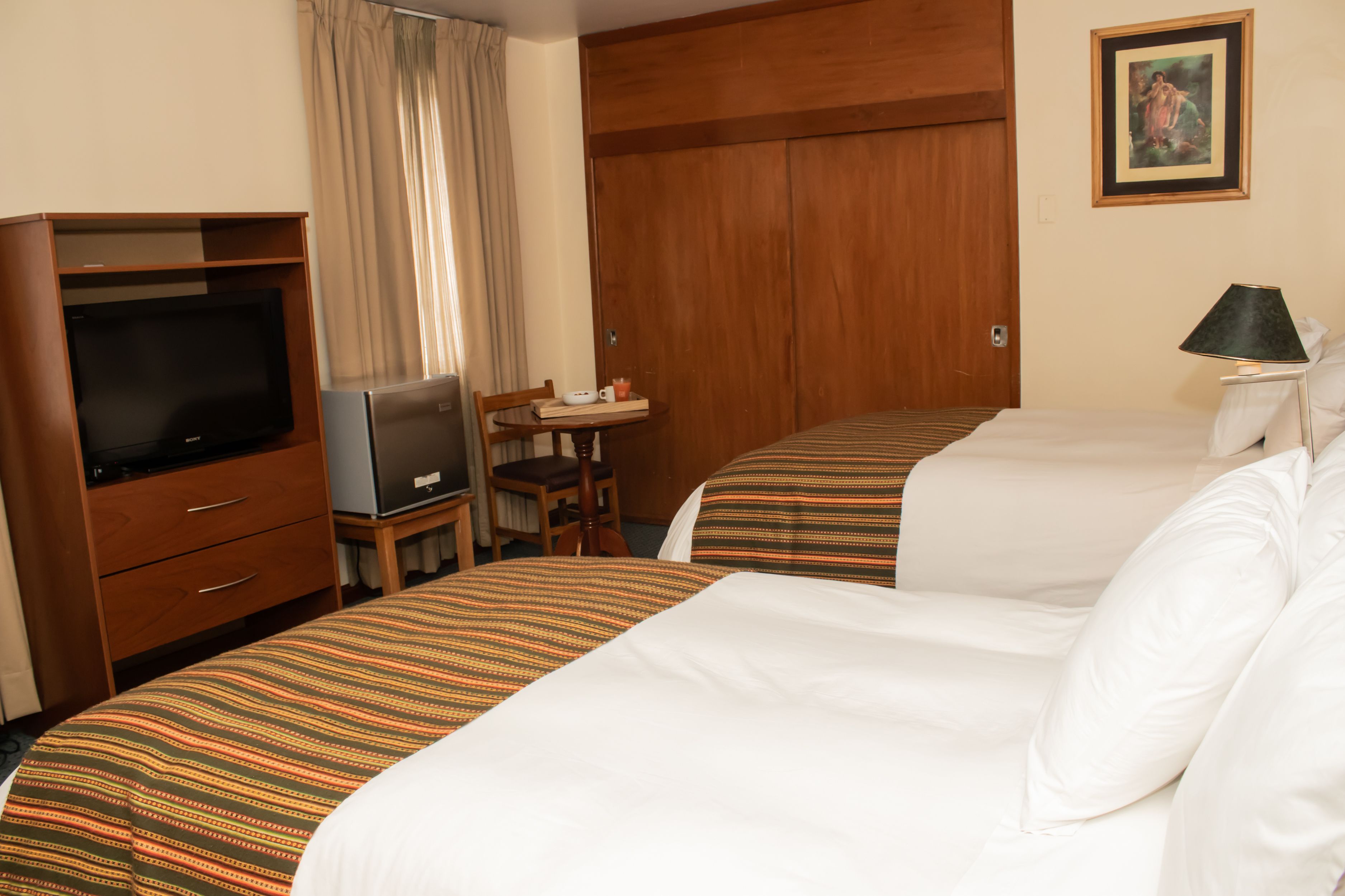 A hotel room with two beds, a TV, a desk with a chair, and a lamp.