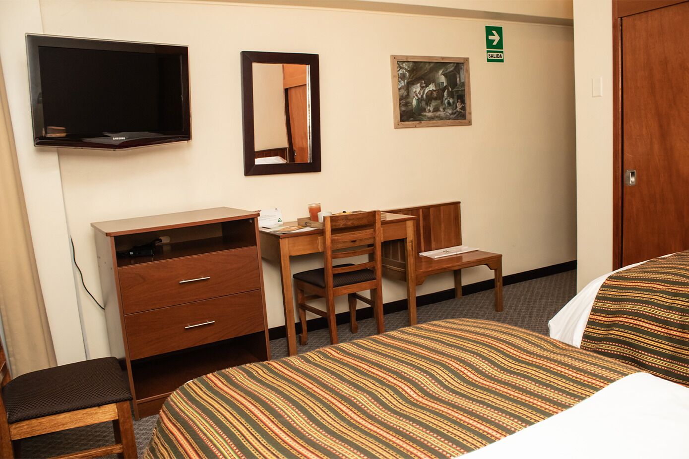A hotel room with a bed, a desk, a chair, a dresser, a TV, and a painting on the wall.
