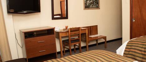 A hotel room with a bed, a desk, a chair, a dresser, a TV, and a painting on the wall.