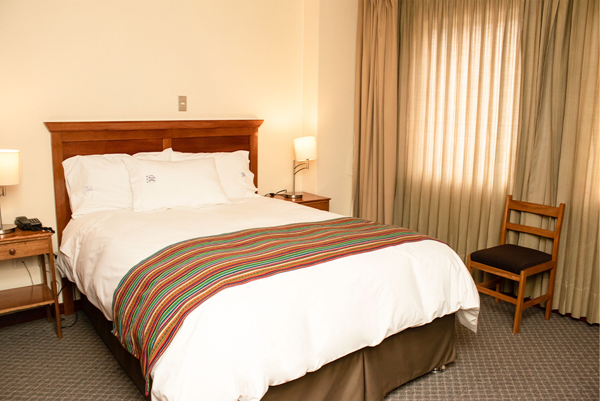 Habitación de hotel con una cama grande, cabecera de madera, mesita de noche con lámpara, una silla y ventana con cortinas.