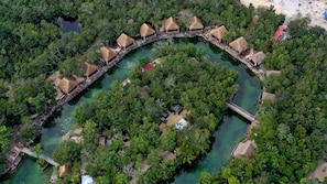 Vista aérea de un resort con bungalows de techo de paja ubicados a lo largo de un canal curvo, rodeado de exuberante vegetación.