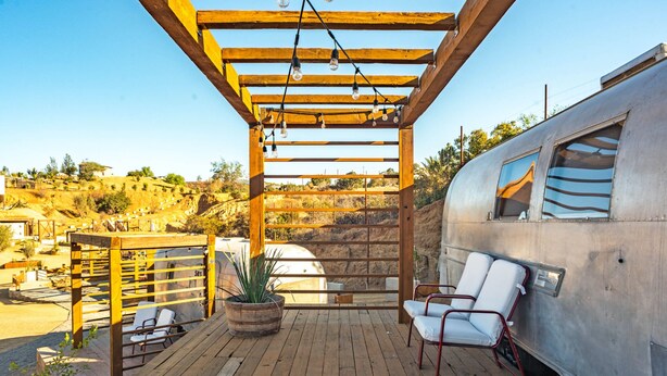Une terrasse en bois avec deux chaises longues blanches, une plante en pot et une pergola décorée de guirlandes lumineuses.