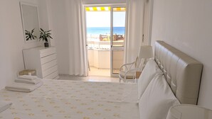 Ein Hotelzimmer mit Bett, Sessel, Lampe, Kommode und Blick auf den Strand.