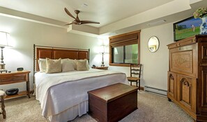 A neatly arranged bedroom with a large bed, a wooden headboard, a bedside table with a lamp, a nightstand, a wooden chest, a dresser, and a television on the wall.