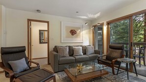 Una sala de estar moderna con un sofá, dos sillones de cuero, una mesa de centro de vidrio y un amplio ventanal con vista a árboles.
