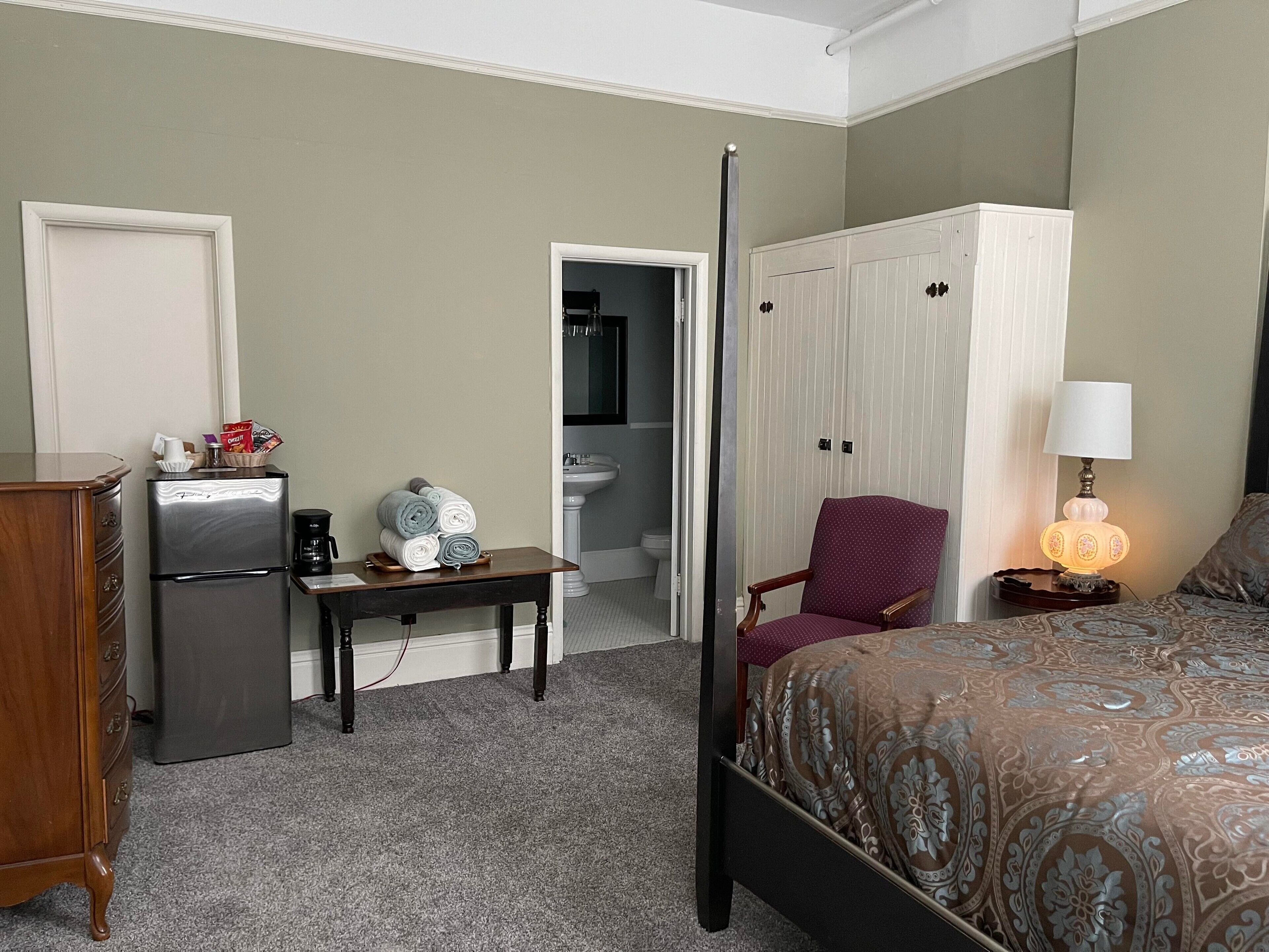 A hotel room with a bed, a desk with a refrigerator, a chair, a bedside table with a lamp, and a wardrobe.