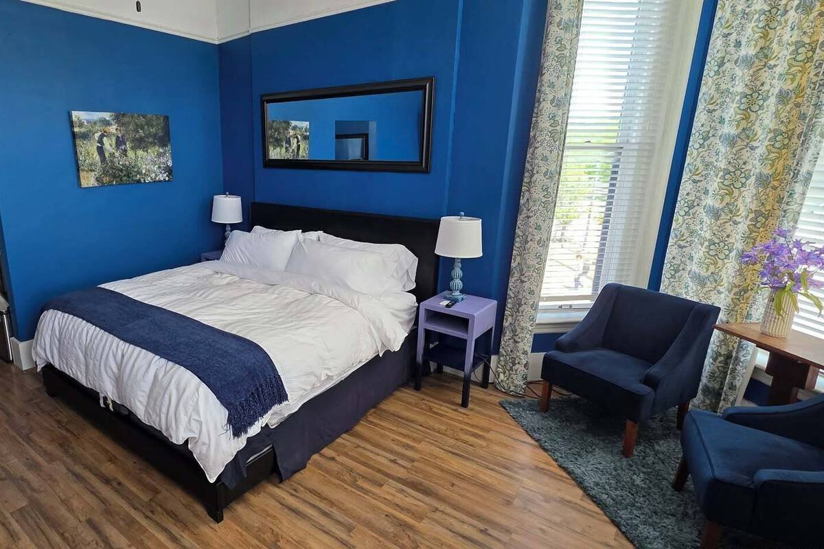 A hotel room with a bed, bedside tables, a chair, a desk, a mirror, and a window with curtains.