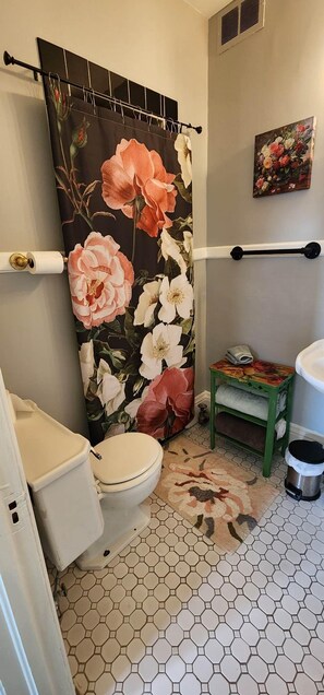 A bathroom with a toilet, a shower curtain with floral patterns, a small table with towels, and a hexagonal tiled floor. - The Clinton Inn (Clinton)