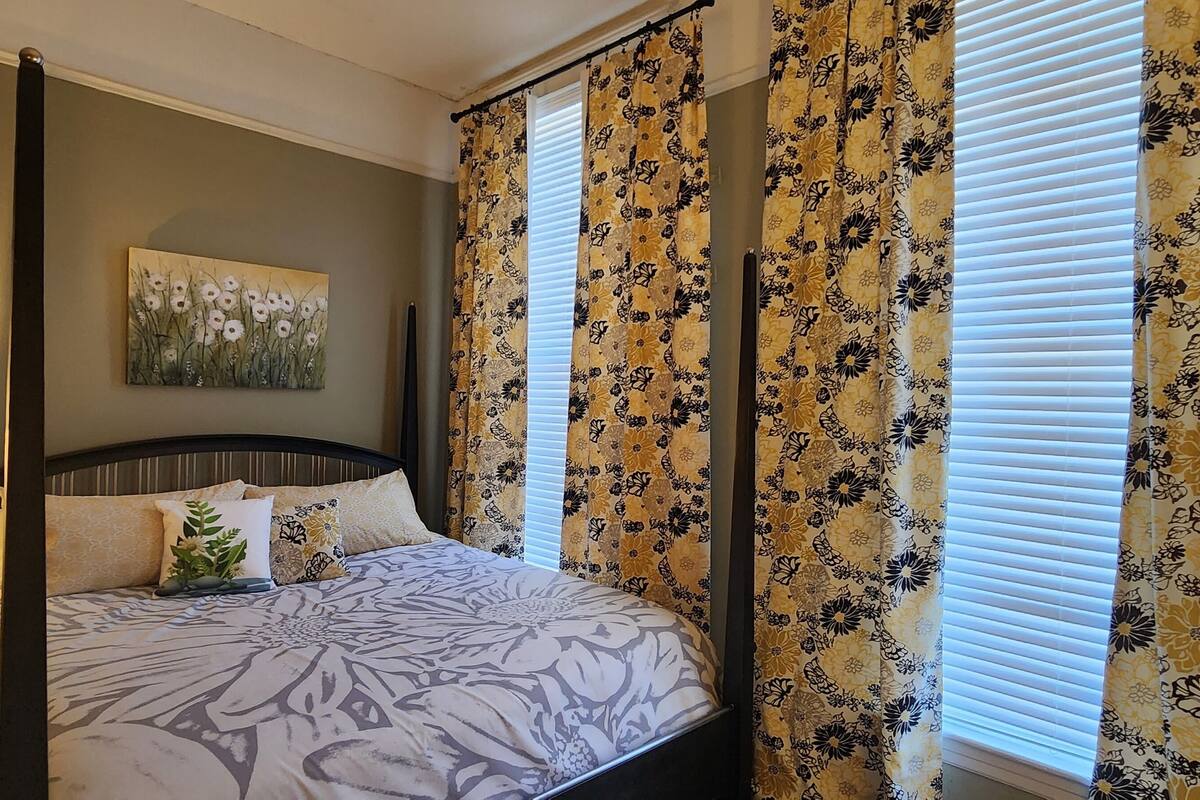 A four-poster bed with a floral bedspread, a nightstand with a lamp, and a painting on the wall.