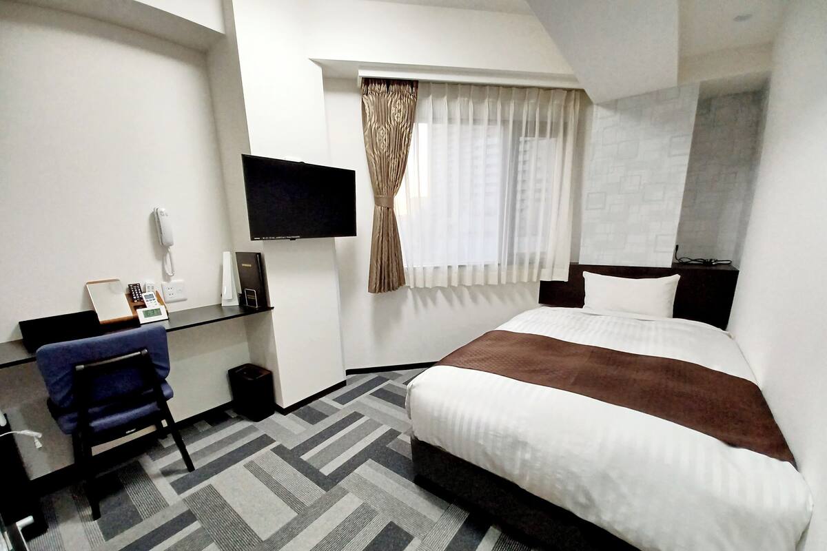 A hotel room with a bed, a desk with a chair, a TV mounted on the wall, and a window with curtains.