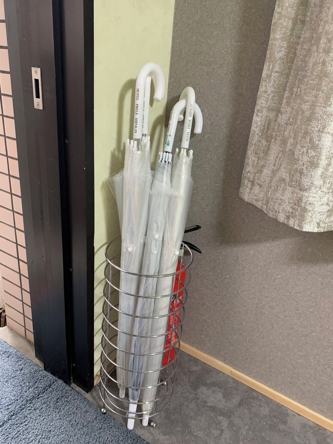 A metal rack holding several white umbrellas with "HOTEL PALLACE SHINKAN" labels, a door, a wall with a curtain, and a red object in the background.