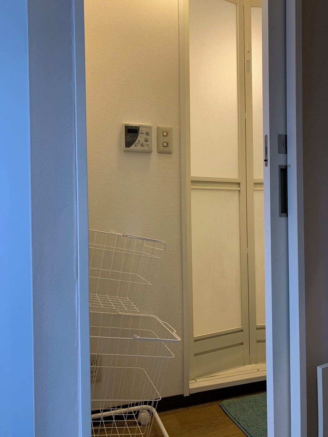 A narrow hallway with a white door, a wall-mounted thermostat, and a white wire basket.