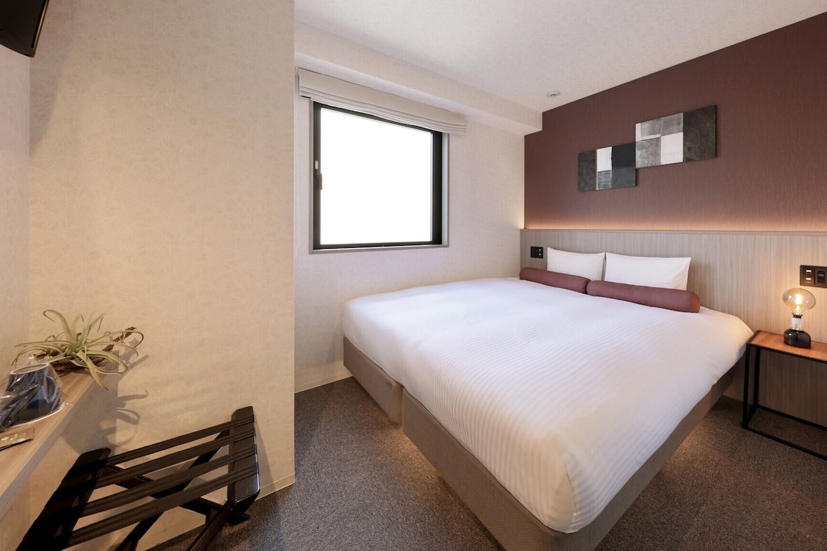 Una habitación de hotel con cama, mesita de noche, lámpara y ventana.