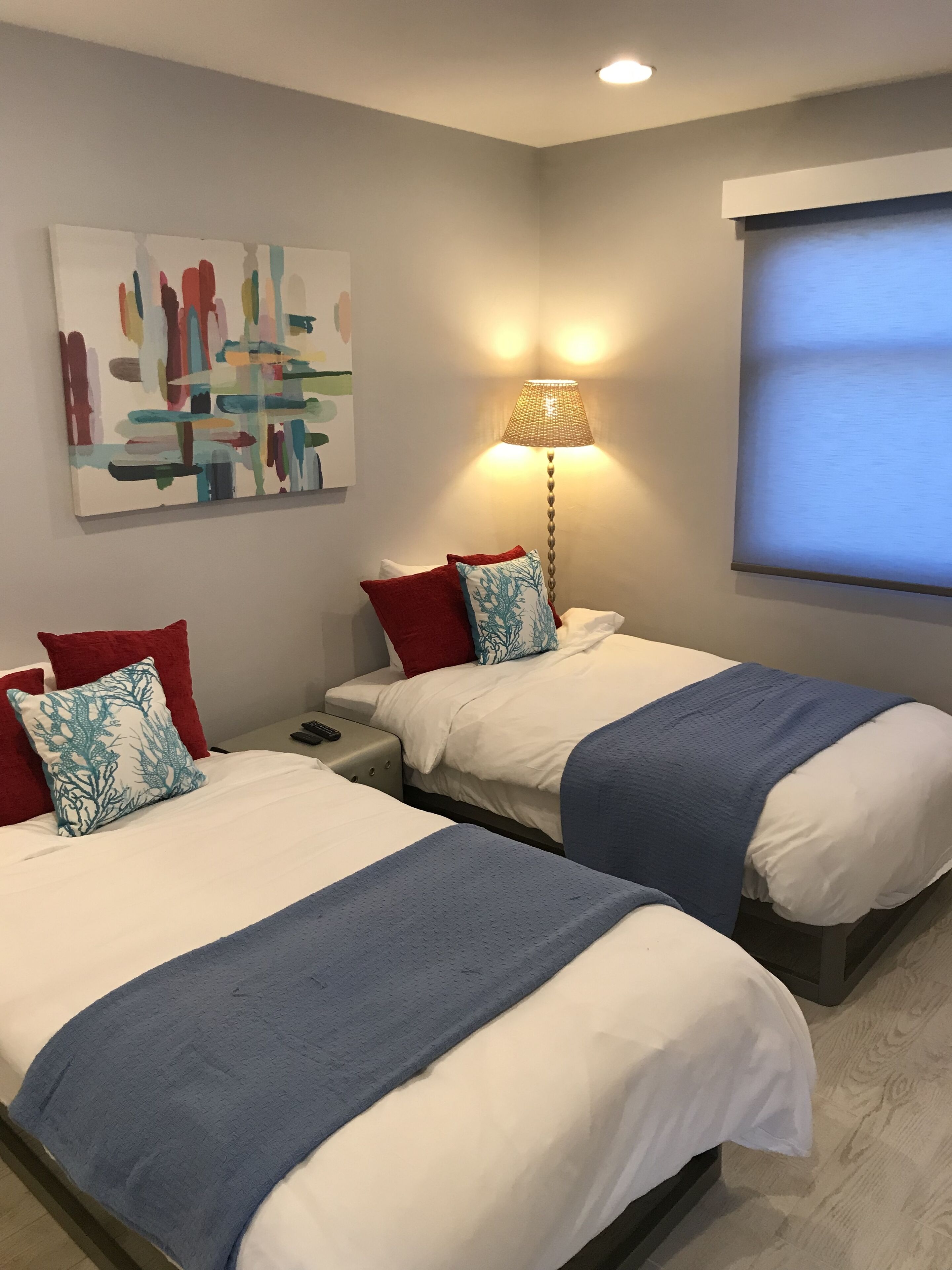 Habitación de hotel con dos camas, cada una con una manta azul y almohadas rojas, una lámpara y un cuadro en la pared.
