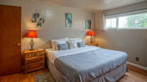 Quarto com cama, duas mesinhas de cabeceira com abajures, porta de madeira e obras de arte nas paredes.