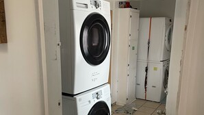 Uma lavanderia com máquinas de lavar e secadoras empilhadas, piso cerâmico e uma porta que dá para outro cômodo.