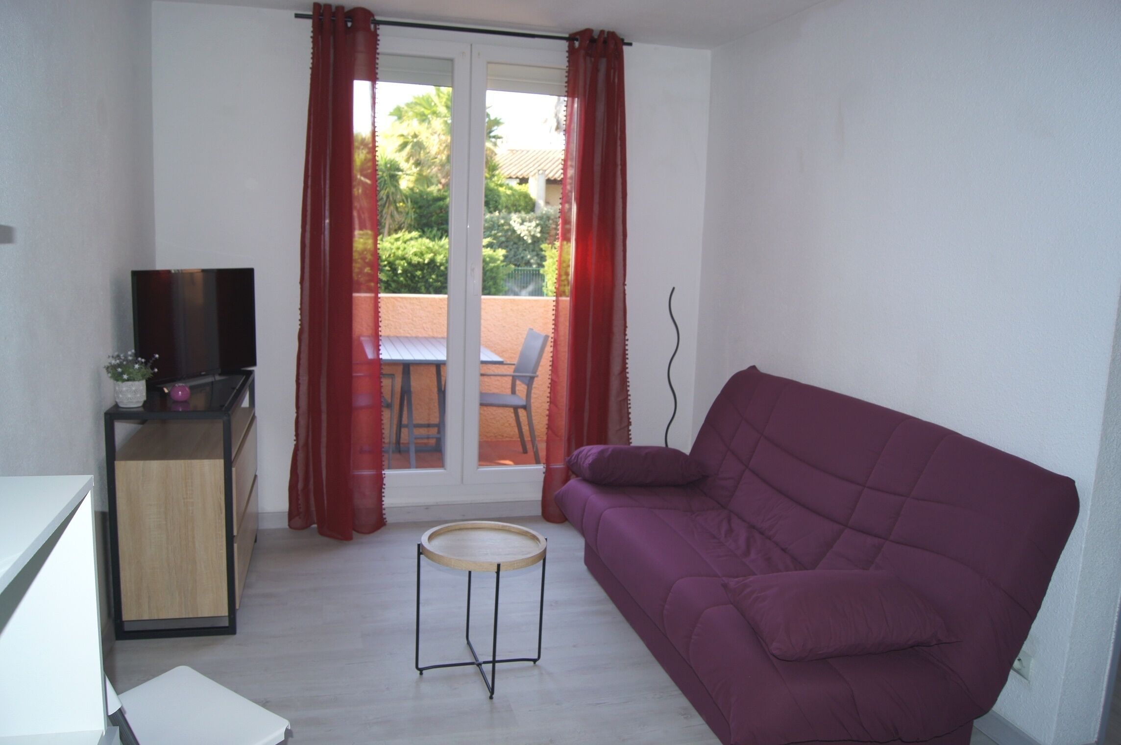 A compact living space with a purple sofa, a small round table, a TV on a stand, and a sliding glass door leading to an outdoor area with a table and chairs.