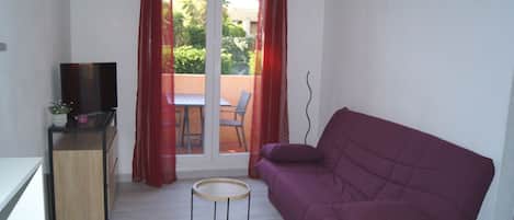 A compact living space with a purple sofa, a small round table, a TV on a stand, and a sliding glass door leading to an outdoor area with a table and chairs.