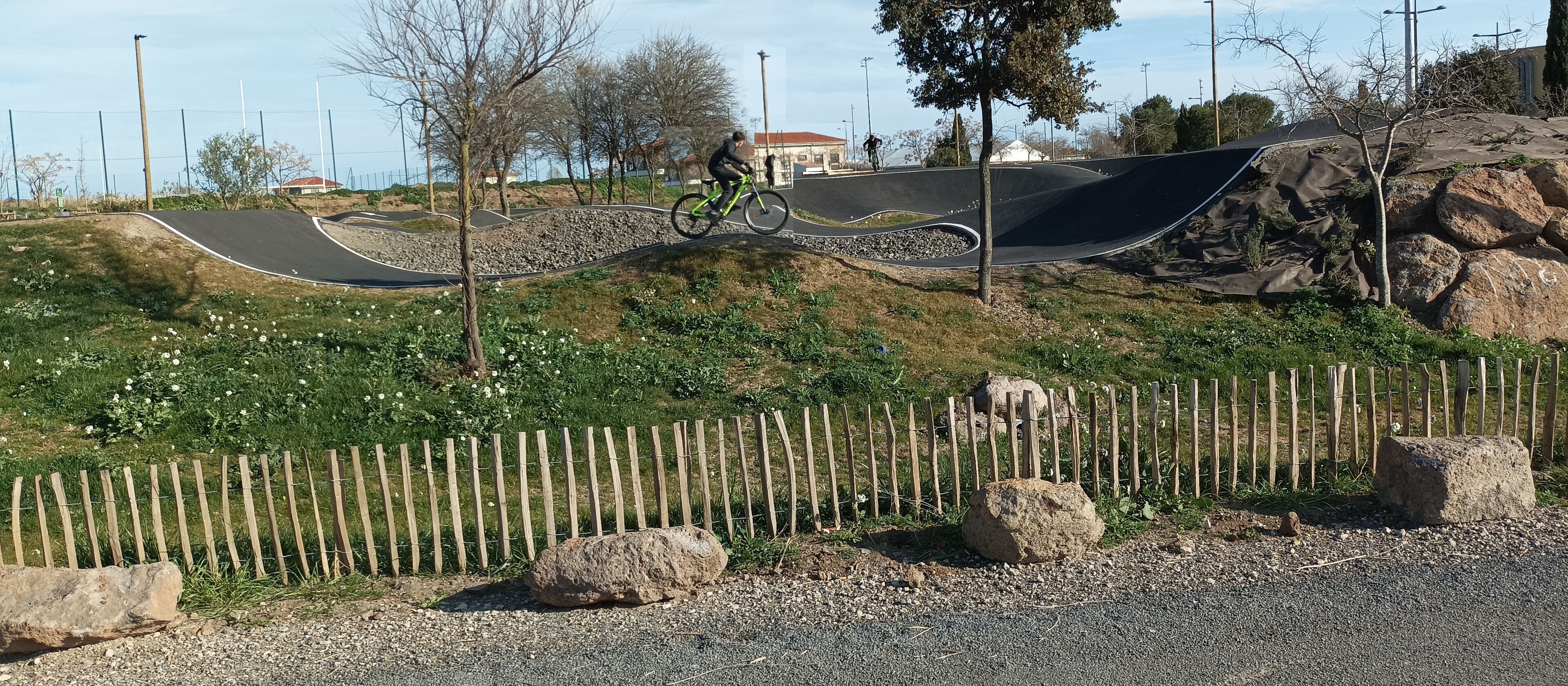 Una pista BMX con ciclistas, rodeada por una valla de madera, rocas y vegetación.