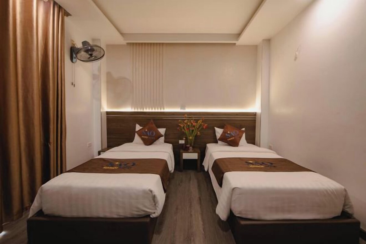 A hotel room with two beds, a nightstand, a wall-mounted lamp, and a window with blinds.