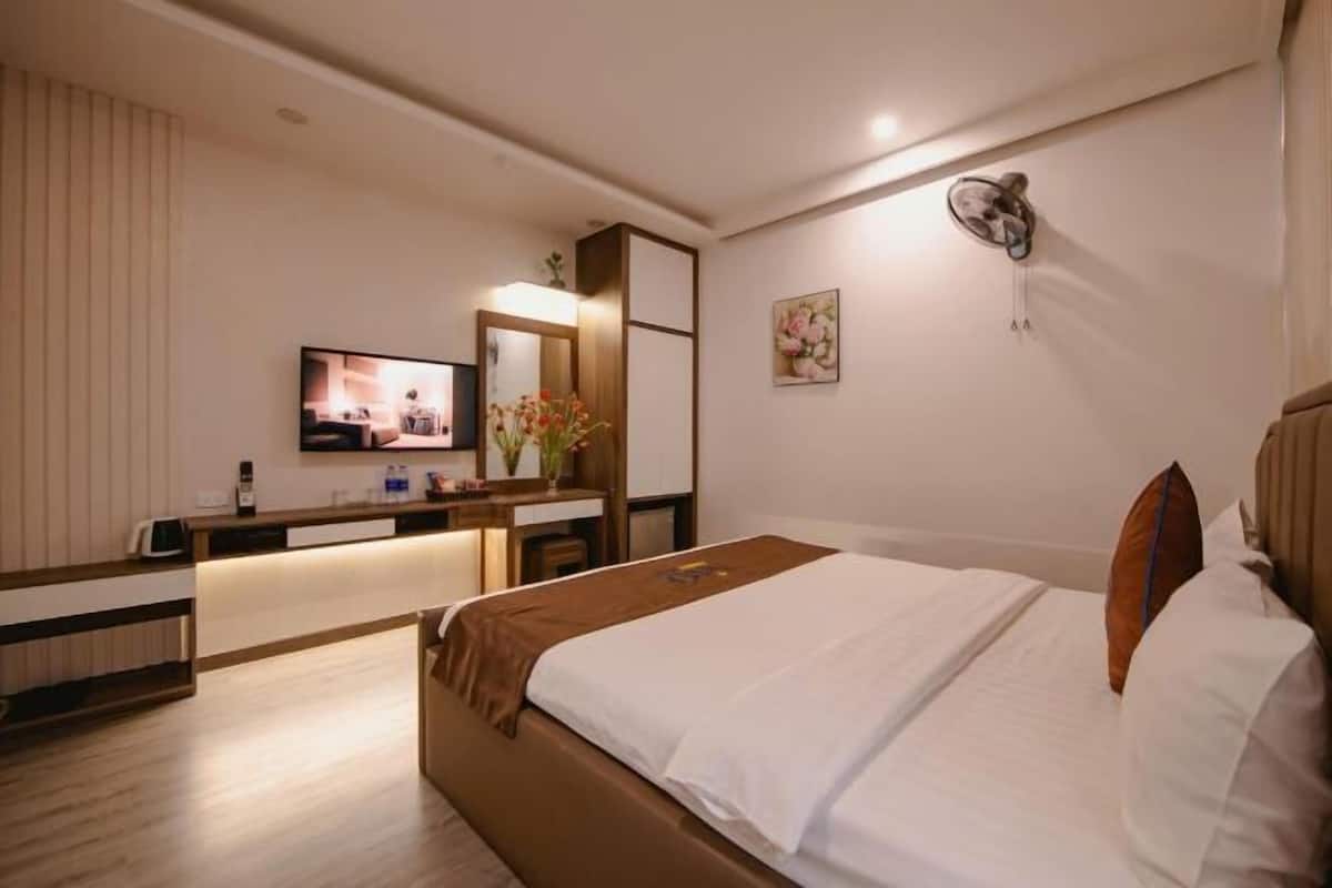 A hotel room with a bed, a desk with a flat-screen TV, a mirror, and a wall-mounted light fixture.