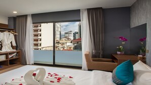 A hotel room with a bed, a TV, a desk with a phone, a wardrobe, and a view of the city. - Orussey One Boutique (Phnom Penh)