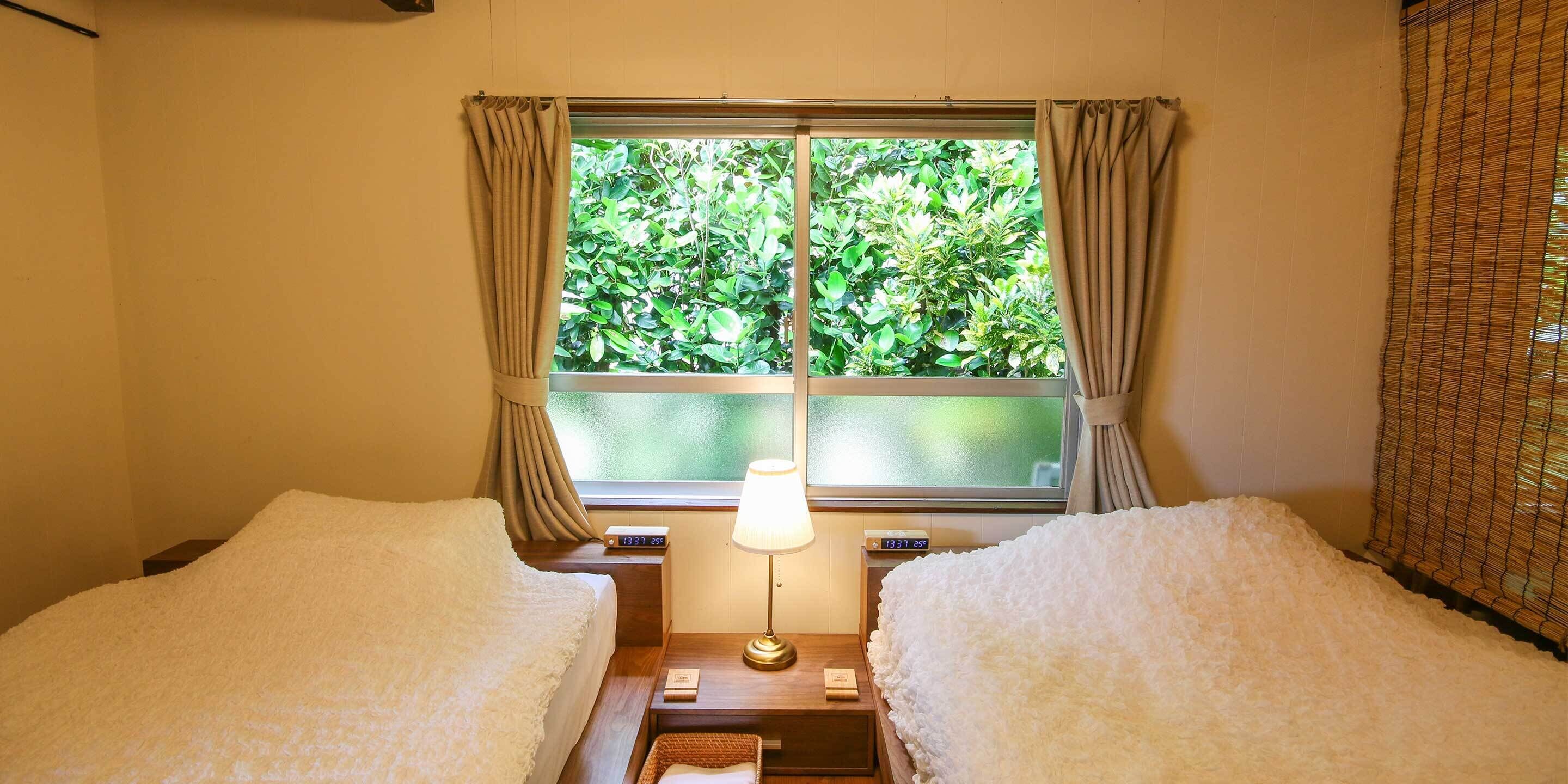 A room with two beds, a window with curtains, a bedside table with a lamp, and a digital clock.