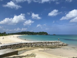 Ein Strand mit einer Steinmauer, klarem türkisblauem Wasser und einer bewaldeten Insel in der Ferne.