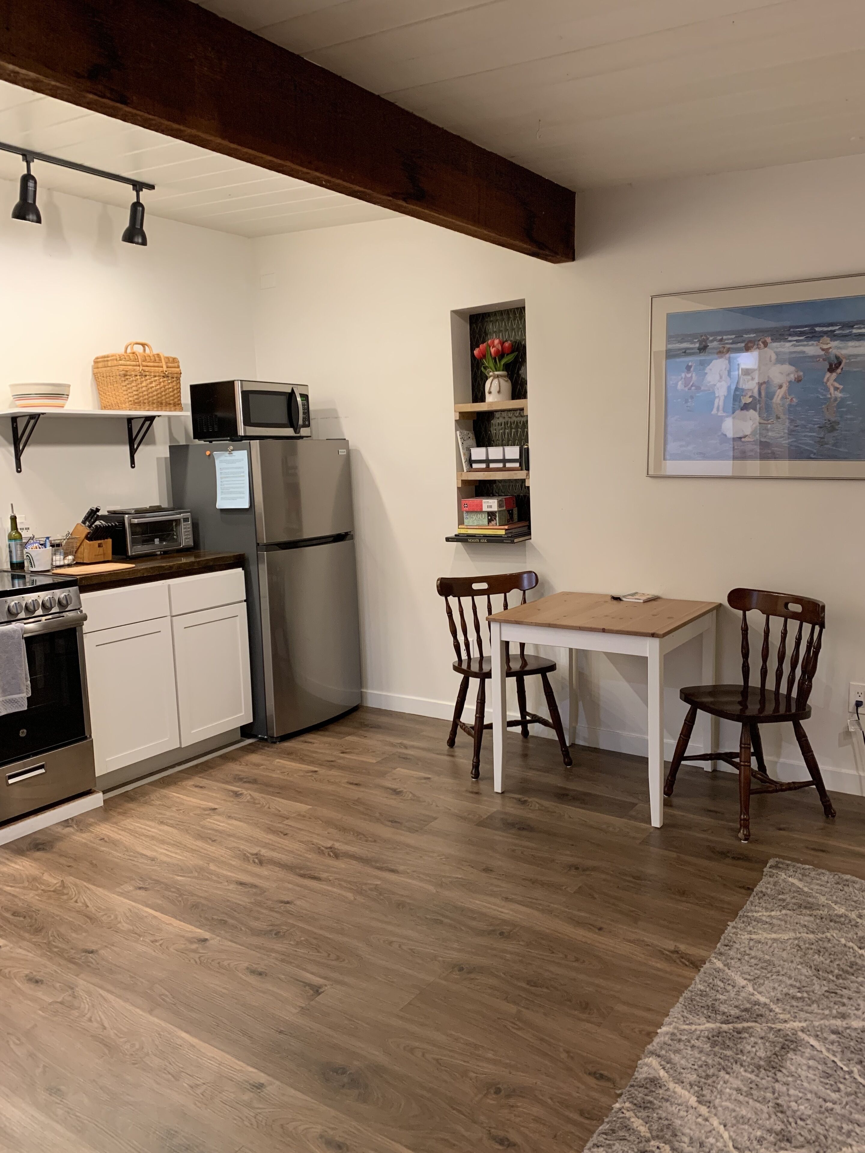 A modern kitchen with white cabinetry, a stainless steel refrigerator, a microwave, and a stove. There is a wooden dining table with chairs and a rug on the floor.