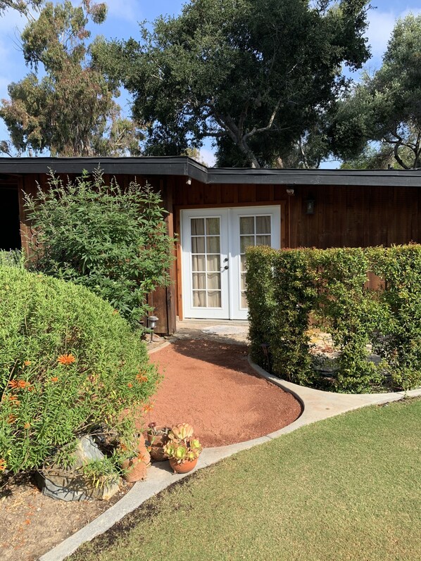 Una casa con puerta principal, un pequeño jardín con diversas plantas y un camino de entrada pavimentado.