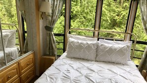 Een net opgemaakt bed met een witte deken, een houten nachtkastje, een spiegel en een raam met uitzicht op groene bomen.