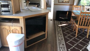 Een compacte keuken met een magnetron, broodrooster en spoelbak. Er is een eethoek met een tafel en stoelen, en een vloerkleed met een patroon.