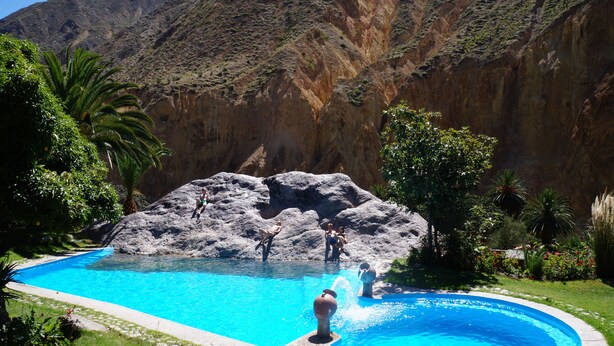 A swimming pool with a rocky island in the center, surrounded by lush greenery and mountains in the background. - Paraiso Las Palmeras Lodge (Cabanaconde)