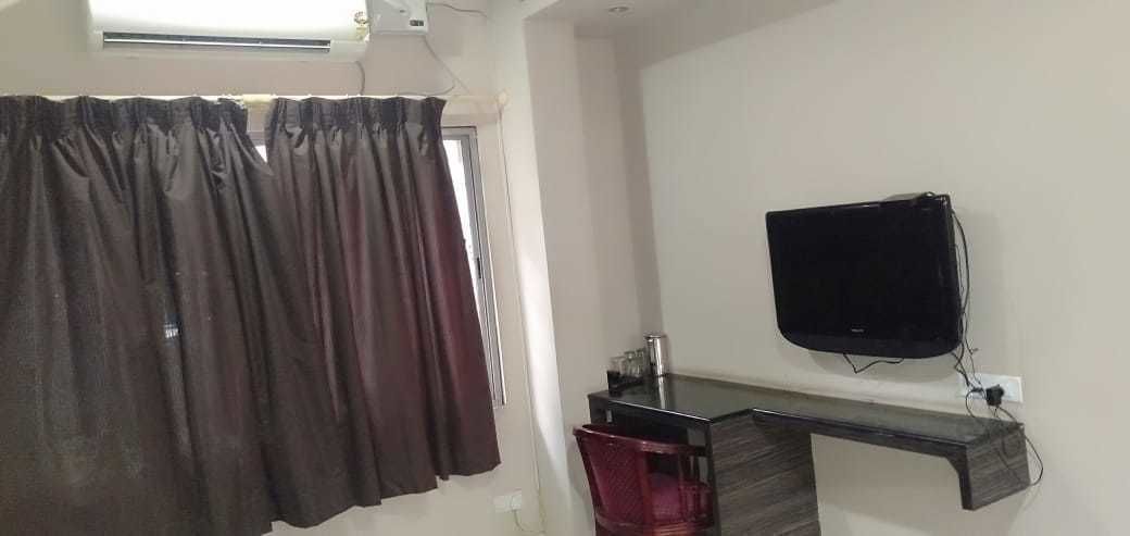 A room with a window covered by dark curtains, a flat-screen TV mounted on the wall, and a small table with a red chair.