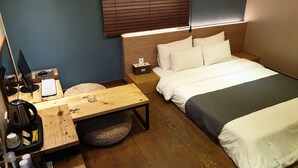 A hotel room with a bed, bedside table, desk with a computer, and a window with blinds.