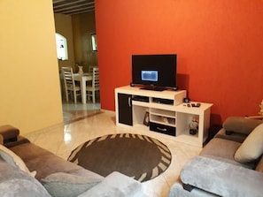 Un salon avec un téléviseur posé sur un meuble, un coin repas à l’arrière-plan et un tapis à motifs.