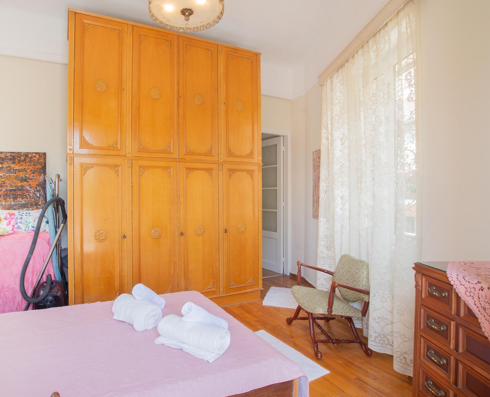 Une chambre avec une grande armoire en bois, un lit avec une literie rose, une chaise et une commode en bois.