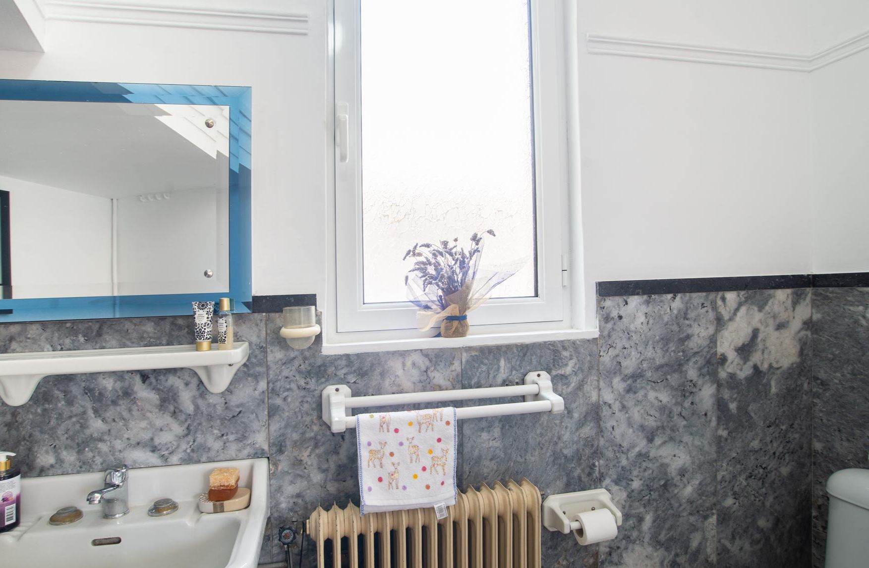 Un bagno con parete in marmo, una finestra vista, un porta asciugamani e uno specchio con cornice blu.