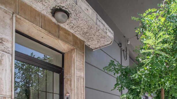 A building entrance with a glass door, a small outdoor area with a tree, and a sidewalk. - Classic Central Athinian Apartment (Athens)