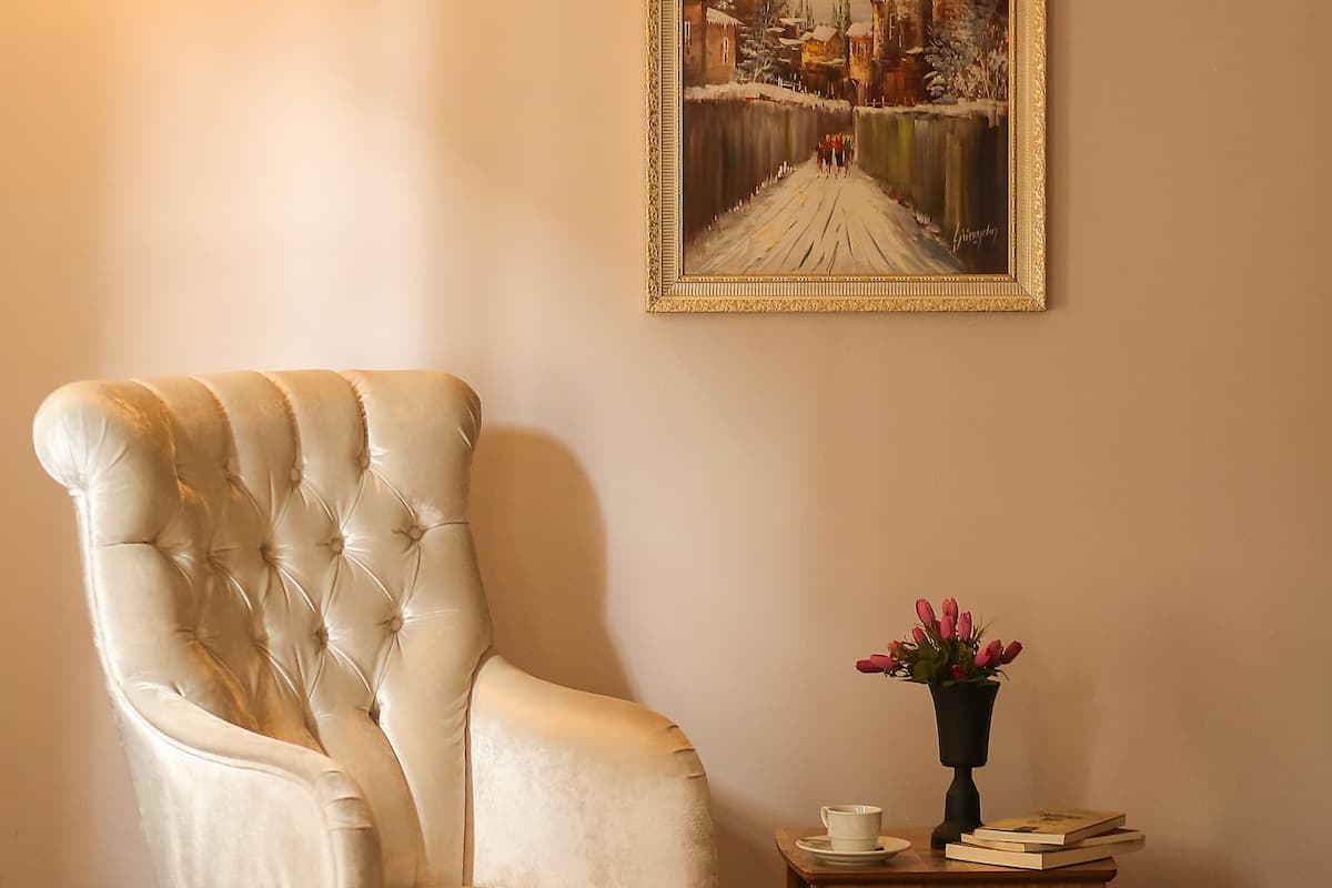 Una habitación con un sillón tapizado, una pequeña mesa de madera con una taza y libros, y un cuadro enmarcado en la pared.