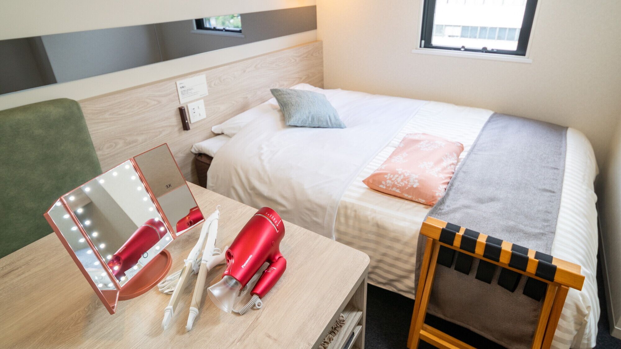 A hotel room with a bed, a bedside table with a mirror and hairdryer, a chair, and a window.