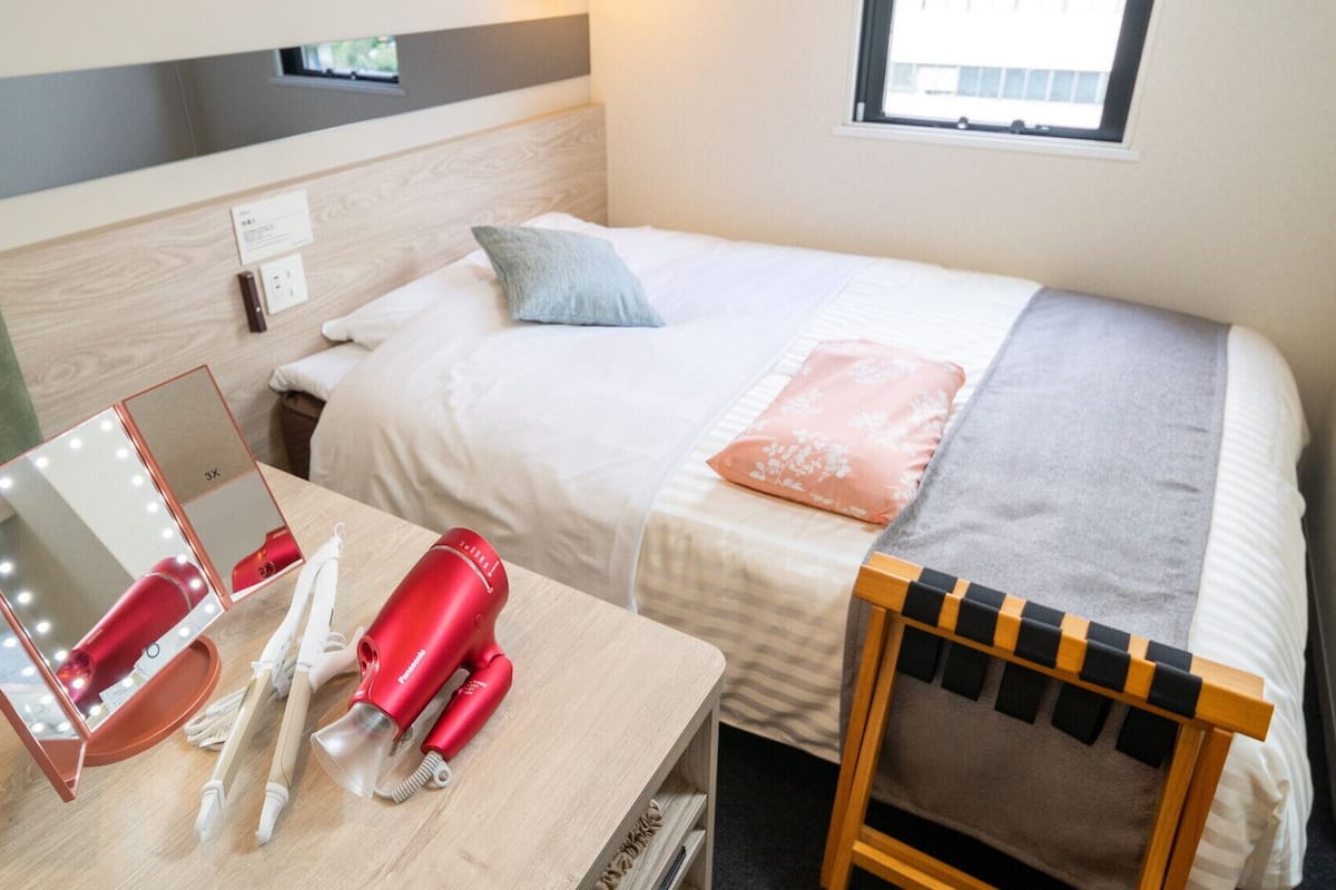 A hotel room with a bed, a bedside table with a mirror and hairdryer, a chair, and a window.