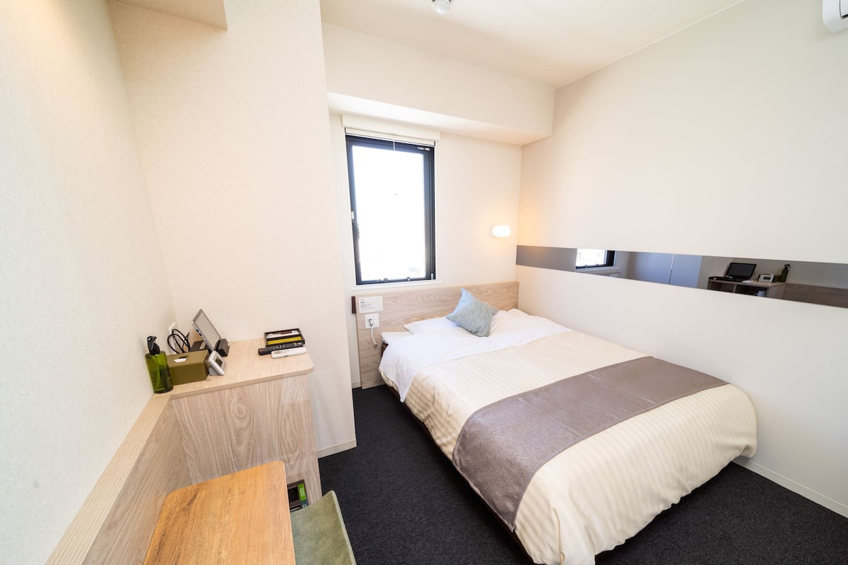 A compact hotel room with a single bed, a small wooden desk, a chair, a bedside table, and a window.